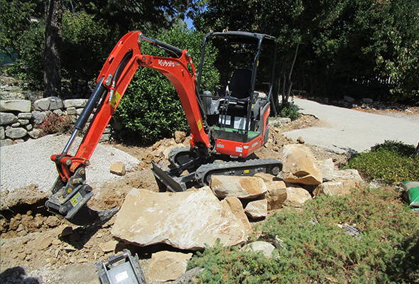 Mini-pelleteuse orange en plein travail de terrassement sur un chantier extérieur, à côté de grandes roches.